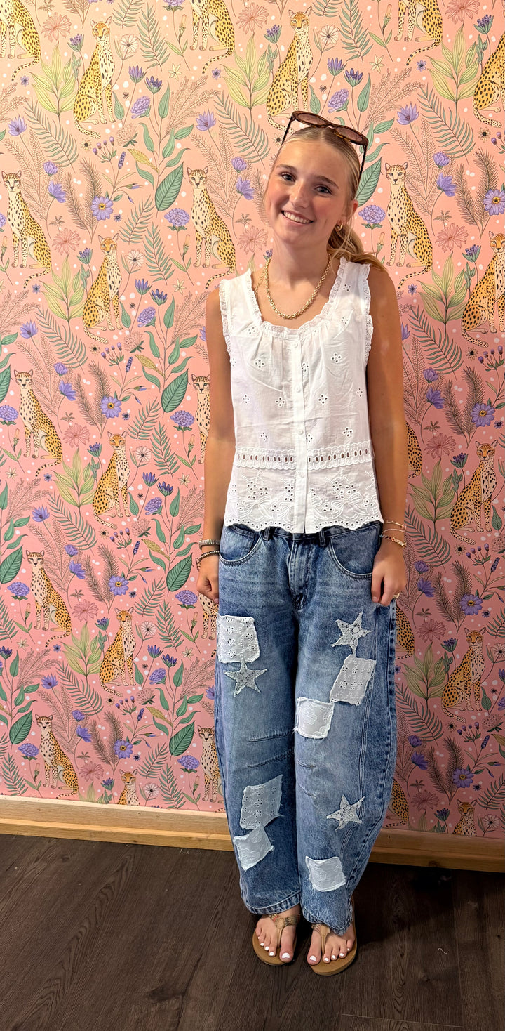 A young woman stands in front of a pink patterned wallpaper background wearing a white sleeveless lace-trim top and blue denim jeans with star patches.