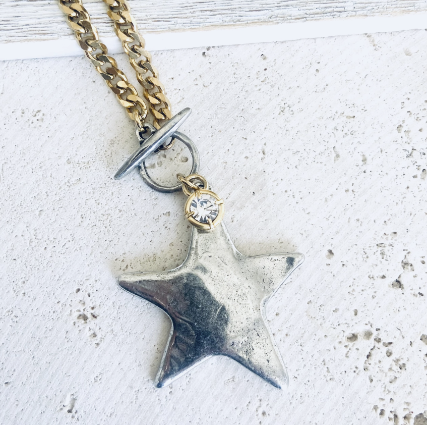 A close-up of a necklace featuring a textured silver star pendant, a small clear rhinestone, and a gold-toned chain with a toggle clasp, resting on a white textured surface.