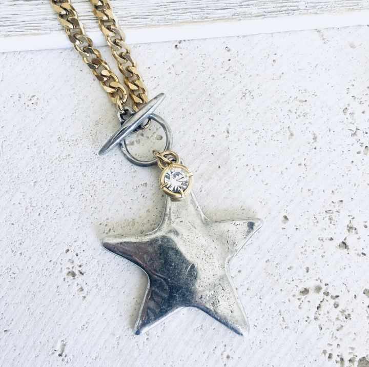 A close-up of a necklace featuring a textured silver star pendant, a small clear rhinestone, and a gold-toned chain with a toggle clasp, resting on a white textured surface.