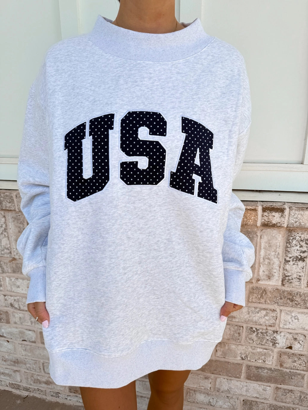 Gray sweatshirt with 'USA' printed on it worn by a person, standing against a brick wall.