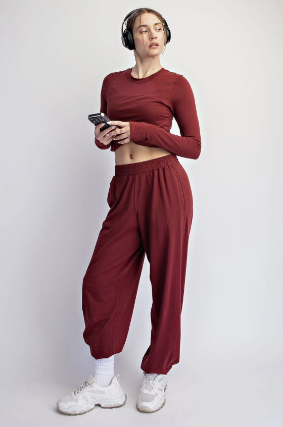 A woman modeling a matching two-piece loungewear set in a deep burgundy red, consisting of a long-sleeve crop top and relaxed sweatpants, paired with white sneakers and headphones against a light gray background.