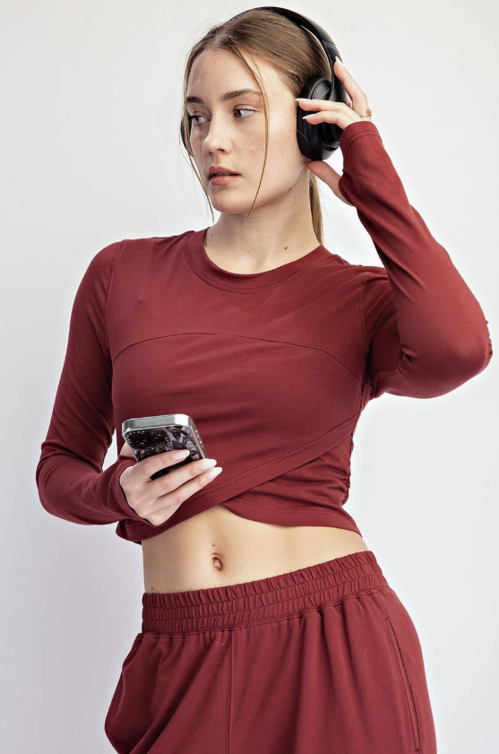 A woman wearing a matching rust-red long-sleeve crop top and sweatpants, holding a smartphone and wearing black over-ear headphones against a light gray studio background.