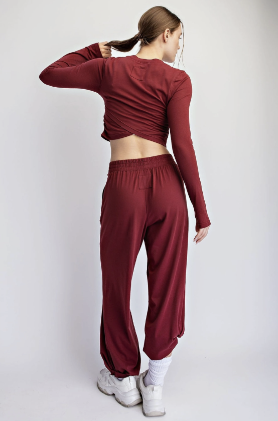 A woman is shown from behind wearing a matching burgundy long-sleeve crop top and jogger pants set, styled with white socks and white sneakers against a light gray studio background.