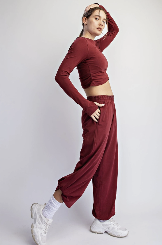 A woman posing in a matching burgundy two-piece activewear set, featuring a long-sleeve crop top and wide-leg sweatpants, styled with white socks and white sneakers against a light gray studio background.