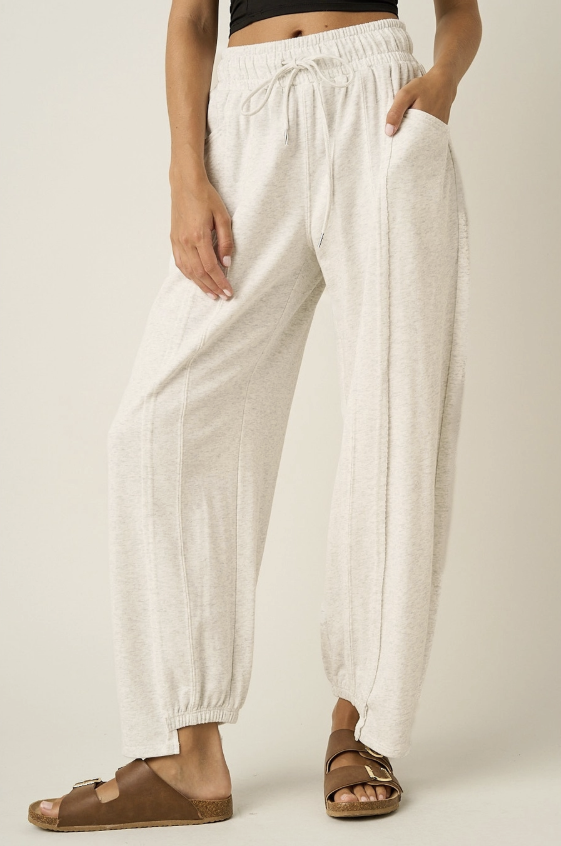 A person wearing loose, drawstring jogger-style pants in a light warm off-white or cream tone, paired with brown double-strap sandals, photographed against a plain light beige studio background.