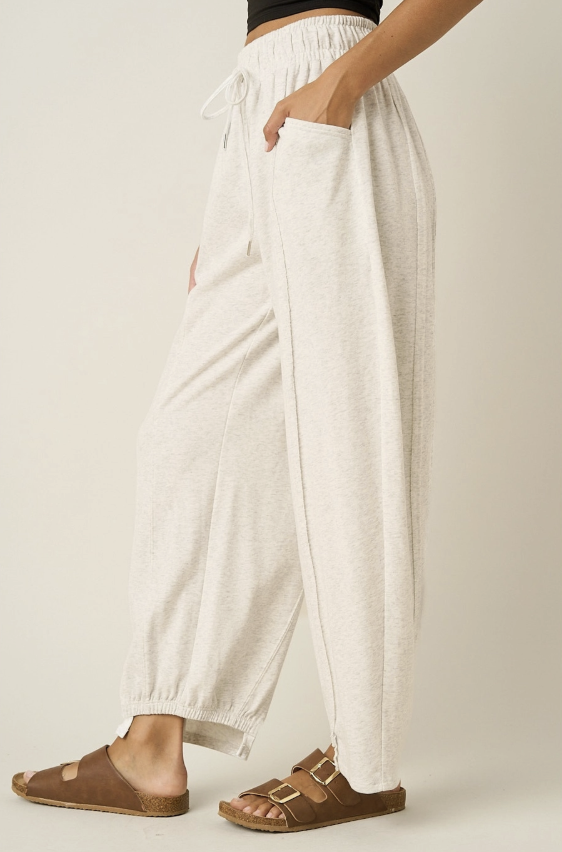 A person wearing loose, light heather cream drawstring lounge pants with side pockets and brown double-strap sandals, shown from the waist down against a plain light beige studio background.