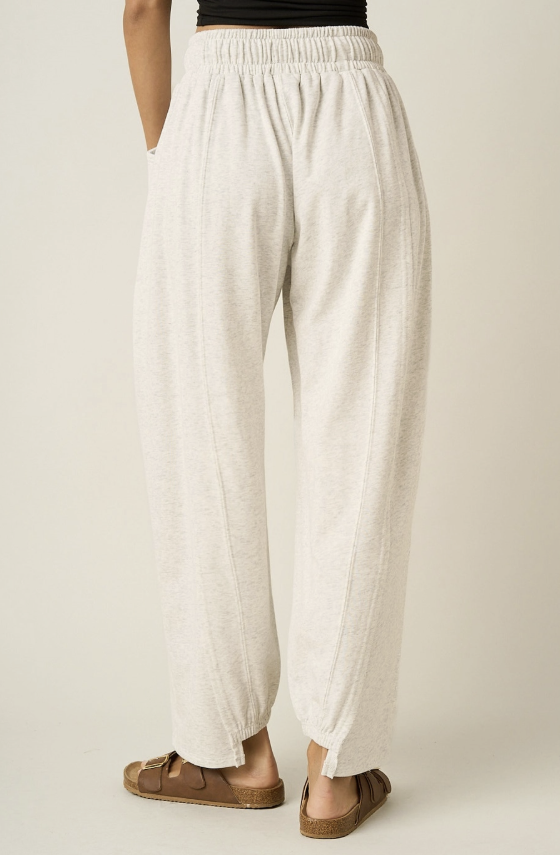 A person shown from the back wearing loose, wide-leg cream or off-white pants with an elastic waistband and brown slide sandals, against a plain light beige studio background.