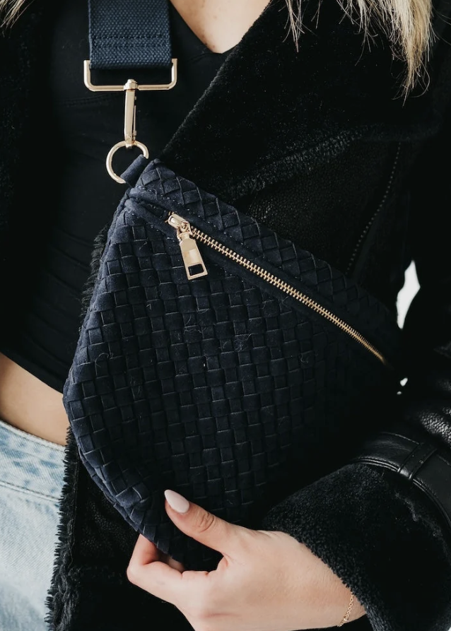 A close-up of a person wearing a black woven crossbody bag with a gold zipper and strap, styled over a black jacket and light blue jeans.