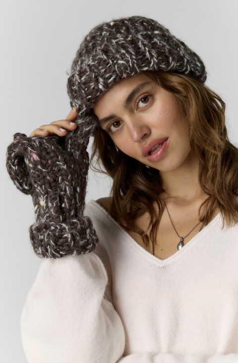 A woman wearing a chunky knit beanie and matching knit mittens in a dark brown and white variegated yarn, posing against a light gray background.
