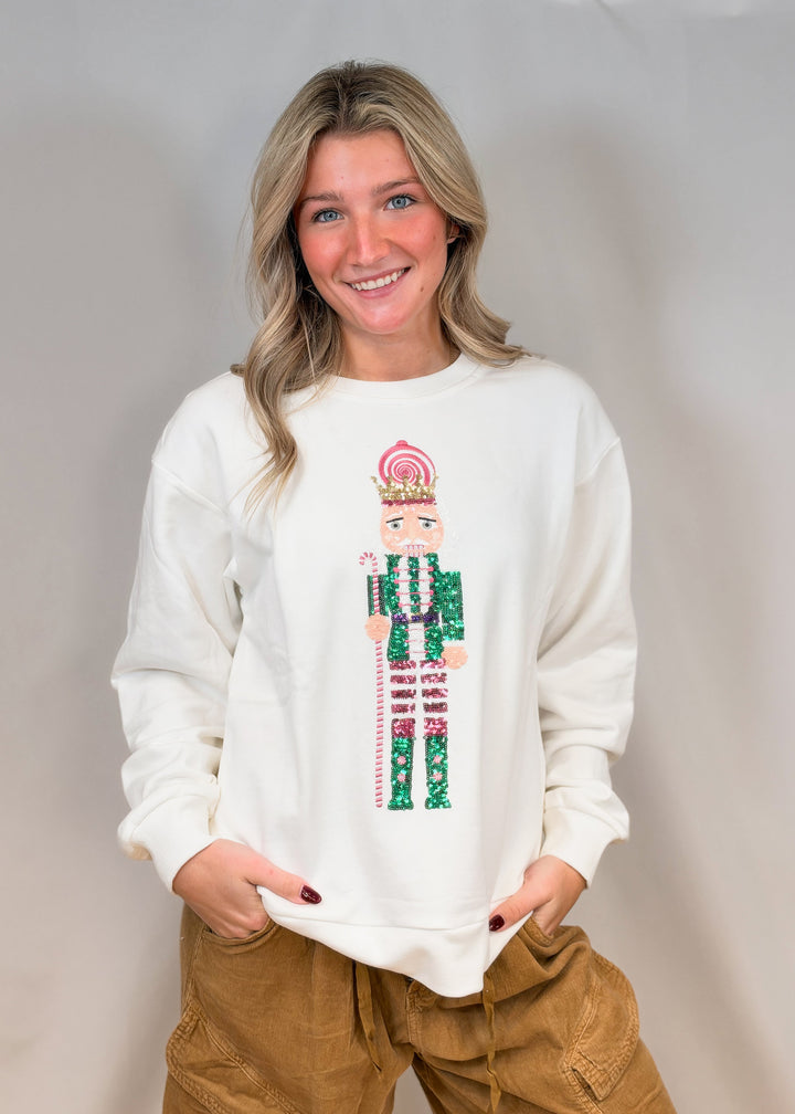 Woman wearing white sweatshirt with sequin nutcracker and brown pants on plain background