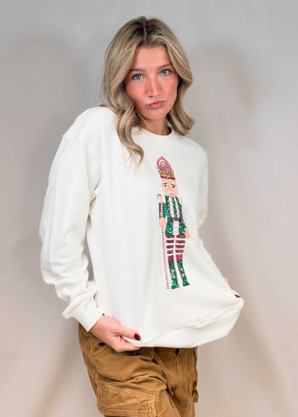 Woman wearing white sweatshirt with sequin nutcracker on front and brown pants on plain background