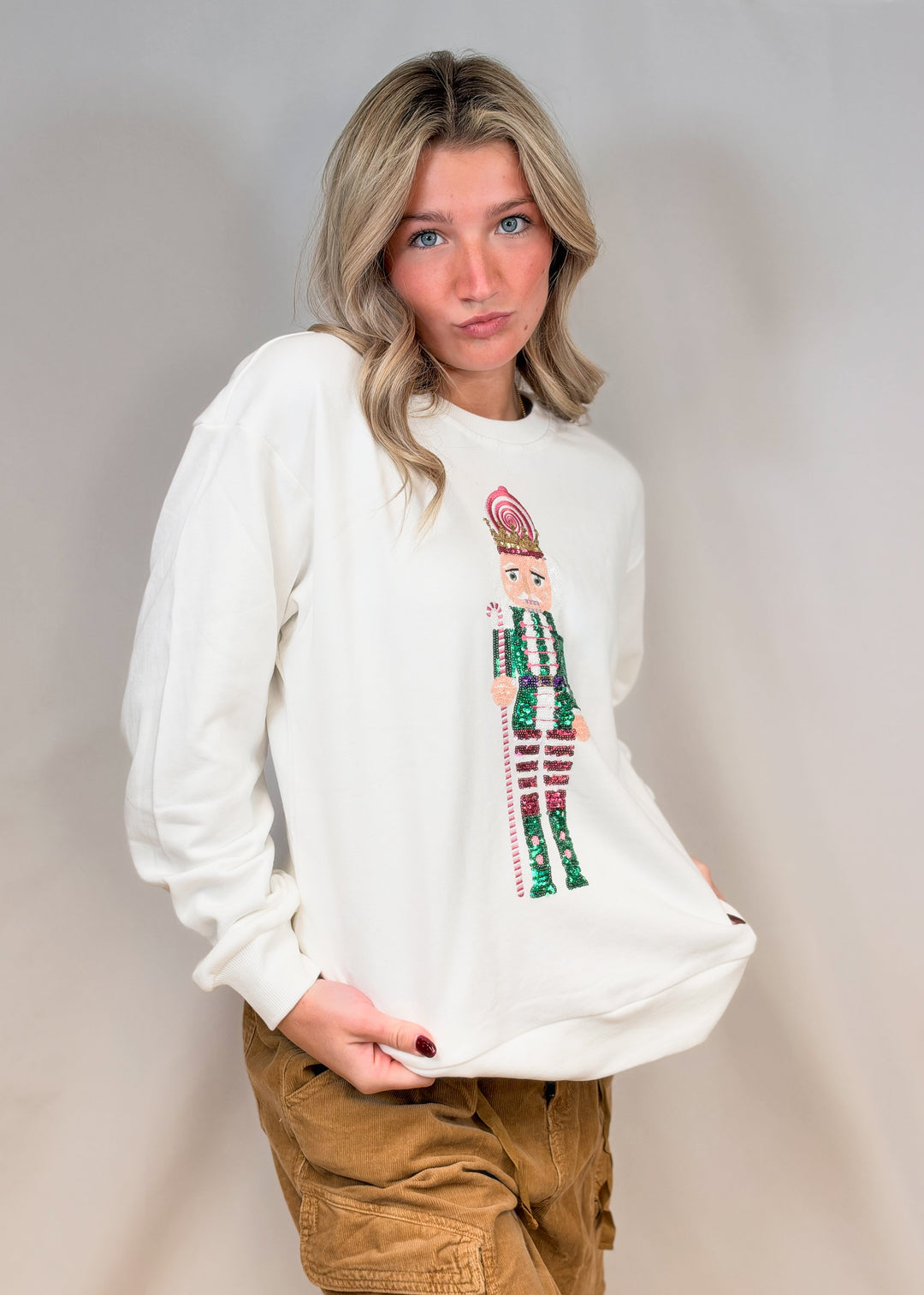 Woman wearing white sweatshirt with sequin nutcracker on front and brown pants on plain background
