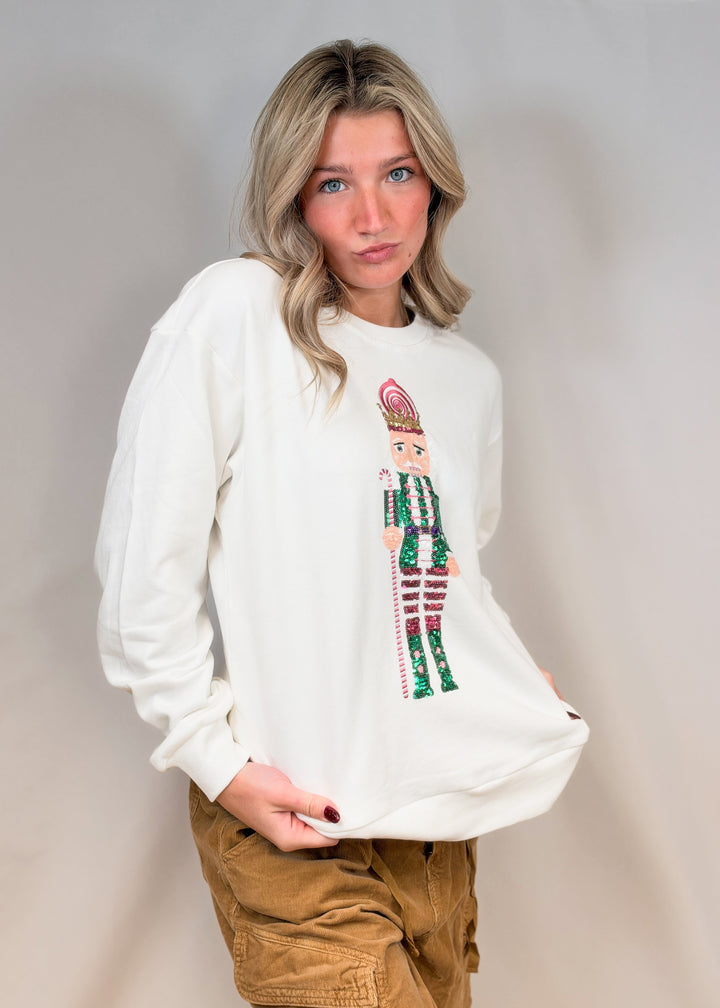 Woman wearing white sweatshirt with sequin nutcracker on front and brown pants on plain background