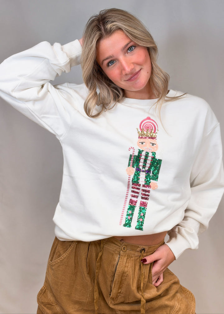 Woman wearing white sweatshirt with sequin nutcracker and brown pants on plain background