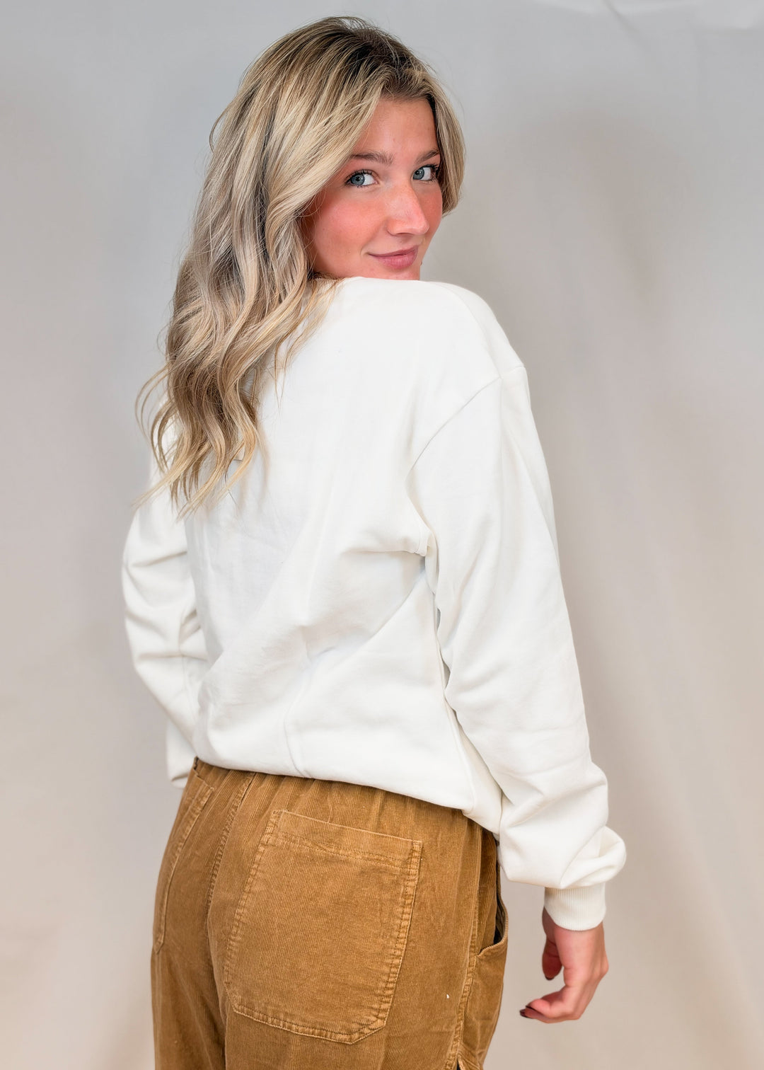 Woman shown from back wearing white sweatshirt and brown pants on plain background