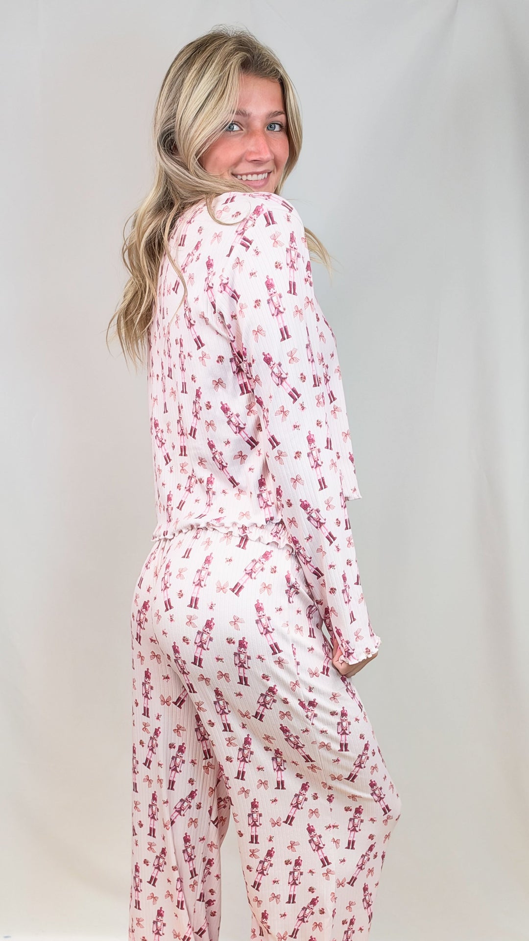 Woman shown from back wearing pink christmas pajamas with nutcrackers and bows on plain background