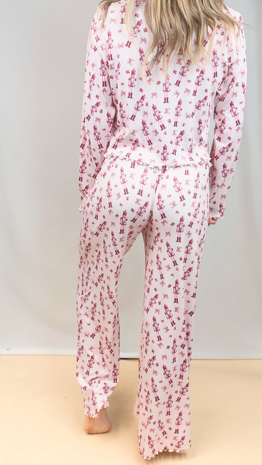 Woman shown from back wearing pink christmas pajamas with nutcrackers and bows on plain background