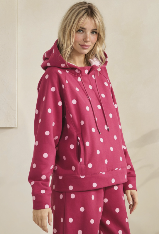 A woman wearing a matching pink and white polka-dot lounge set, featuring a pullover hoodie and coordinating pants, posed against a neutral beige background.