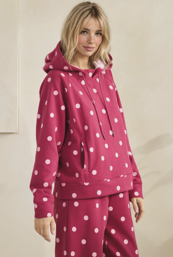 A woman wearing a matching pink and white polka-dot lounge set, featuring a pullover hoodie with drawstrings and matching sweatpants, set against a neutral beige background.