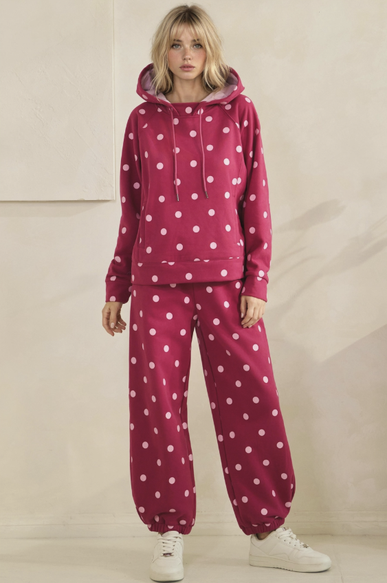 A woman modeling a matching two-piece loungewear set consisting of a hooded sweatshirt and jogger pants, both in a deep magenta pink with a white polka dot pattern, paired with white sneakers.