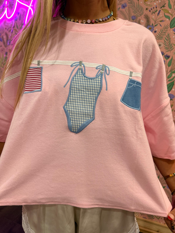 Pink sweatshirt with graphic of a swimsuit and jeans on a patterned background