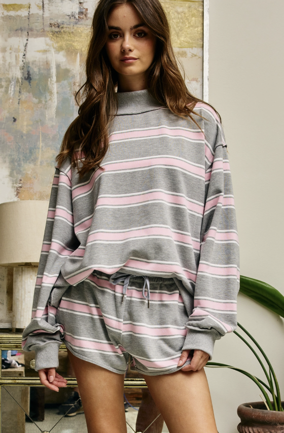 A woman wearing a gray mock-neck sweatshirt and matching drawstring shorts with pink and white horizontal stripes, posed in a styled indoor setting.
