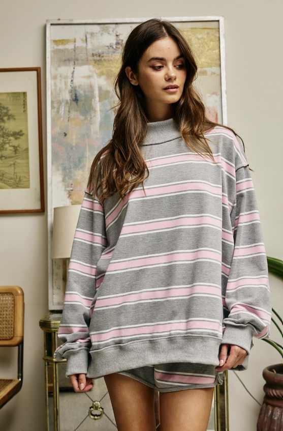 A woman wearing an oversized gray sweatshirt with pink and white horizontal stripes, standing in a styled indoor room with framed wall art, a lamp, and furniture.