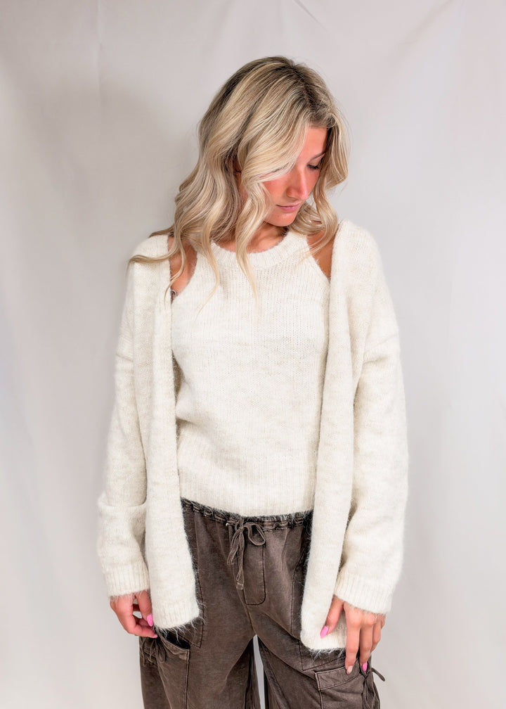 Woman wearing matching cream cardigan and tank with brown pants on plain background