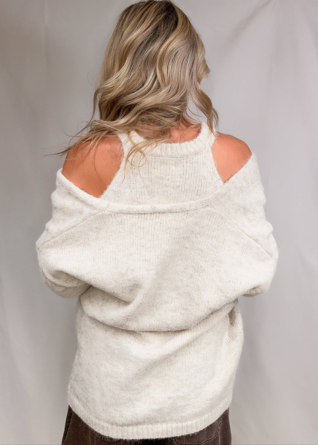 Woman wearing matching cream cardigan and tank with brown pants shown from back on plain background