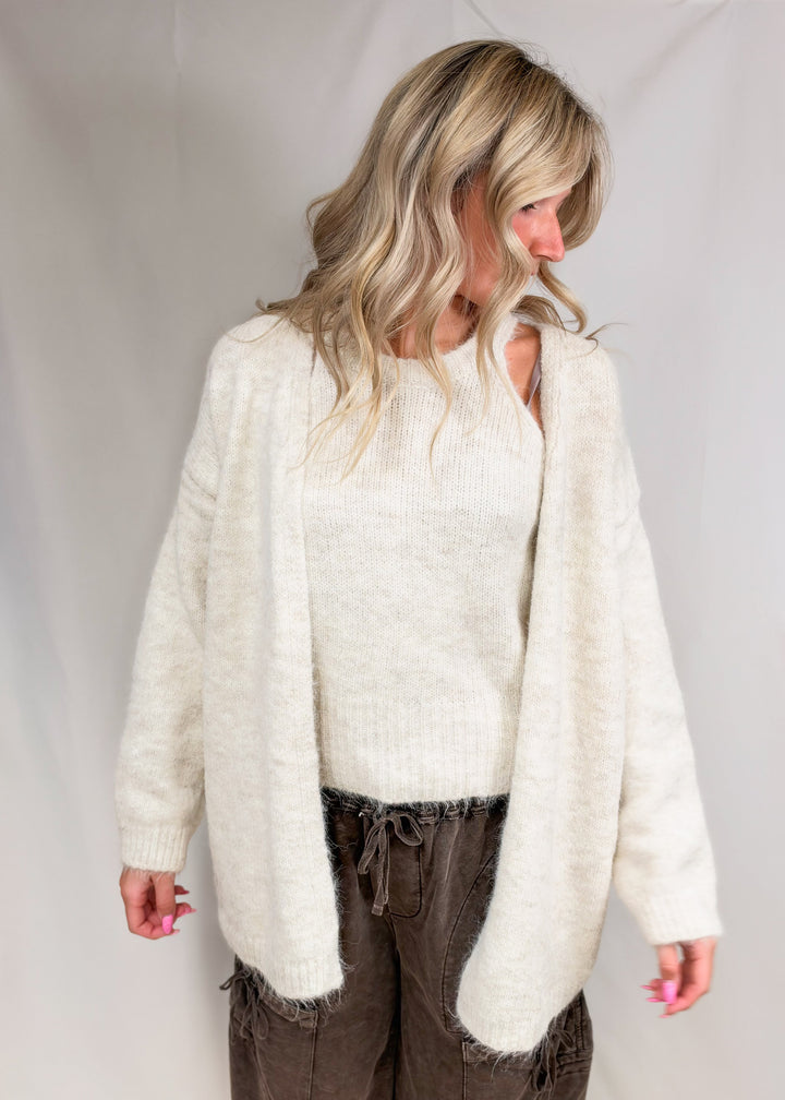 Woman wearing matching cream cardigan and tank with brown pants on plain background