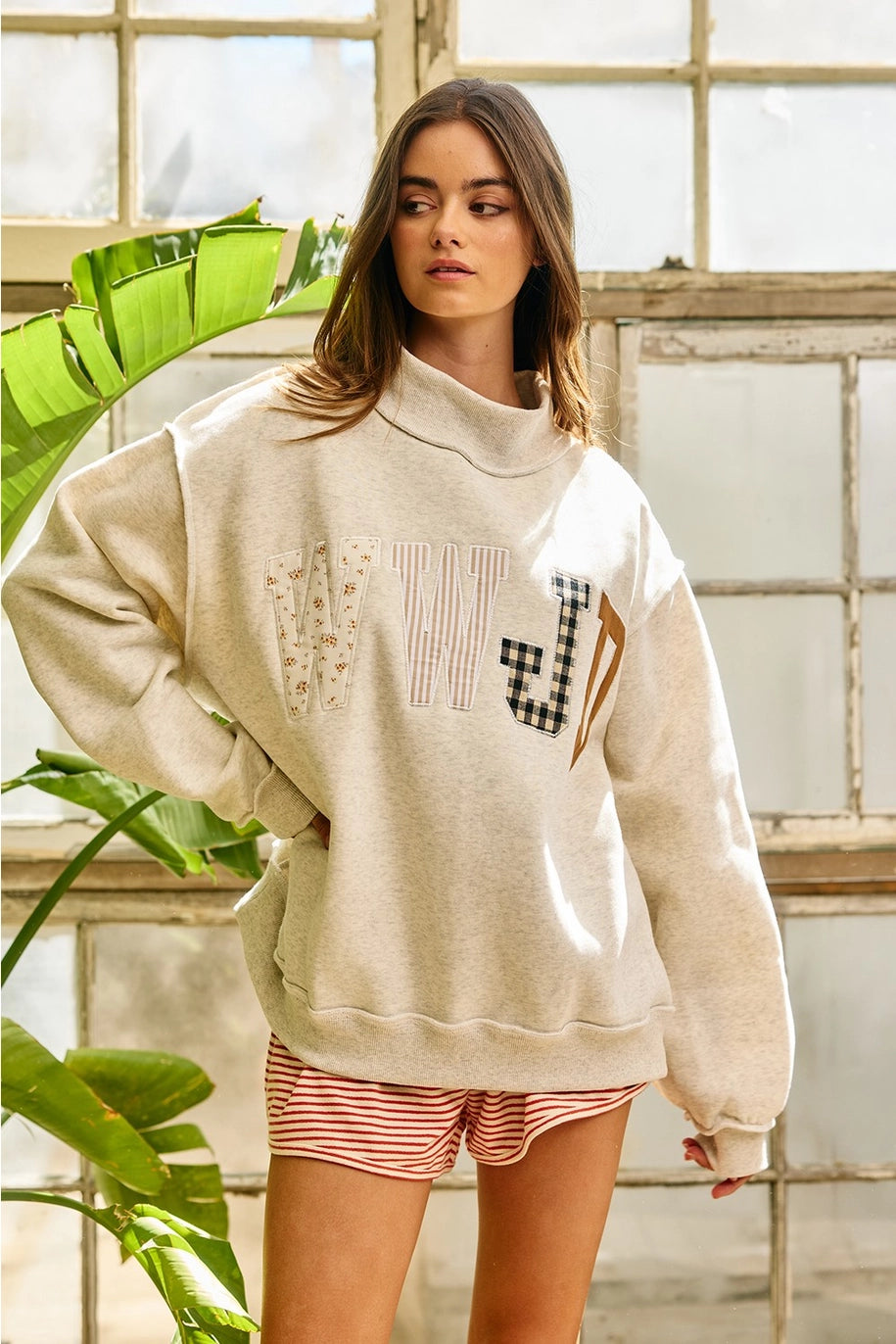 Woman wearing a beige sweatshirt with text and graphics, standing indoors with plants in the background