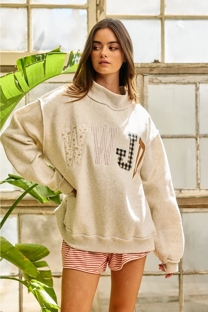 Woman wearing a beige sweatshirt with text and graphics, standing indoors with plants in the background