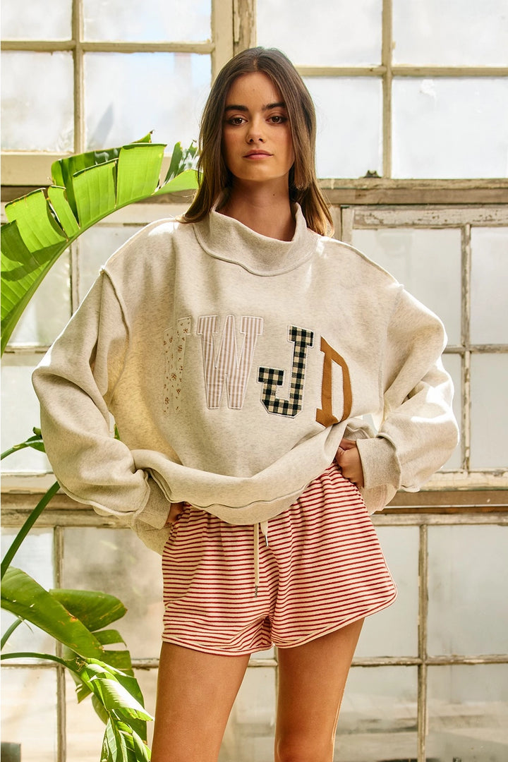 Woman wearing a beige sweatshirt with letter designs and pink shorts in a room with large windows and plants.