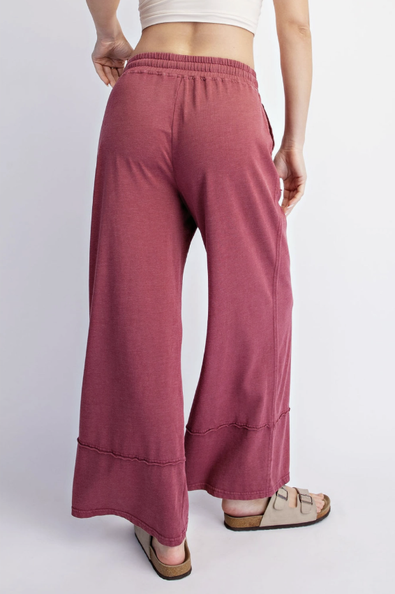 A person shown from the back wearing loose, wide-leg dusty rose pants with an elastic waistband and cuffed hems, paired with beige slide sandals against a plain light gray studio background.