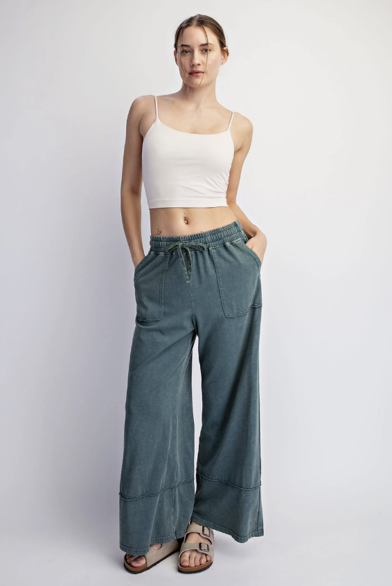 A woman modeling a pair of wide-leg, knee-cuffed pants in a dusty teal-blue color, paired with a white crop top and brown sandals, against a light gray studio background.