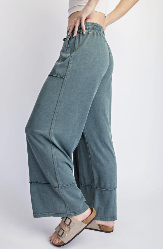 A person wearing wide-leg, cropped wide-leg pants in a muted blue-green denim-like fabric, paired with beige slide sandals, shown against a plain light gray studio background.