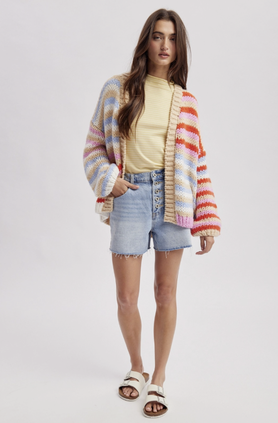 A model stands against a light gray studio background wearing a pastel multicolor striped knit cardigan over a pale yellow ribbed top, light-wash denim shorts, and white slide sandals.