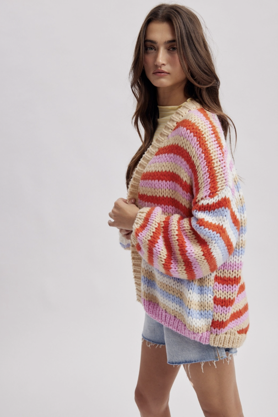 A woman wearing a chunky, multicolor striped knit cardigan sweater with horizontal bands of orange, pink, red, blue, and cream, paired with denim shorts, against a light gray studio background.