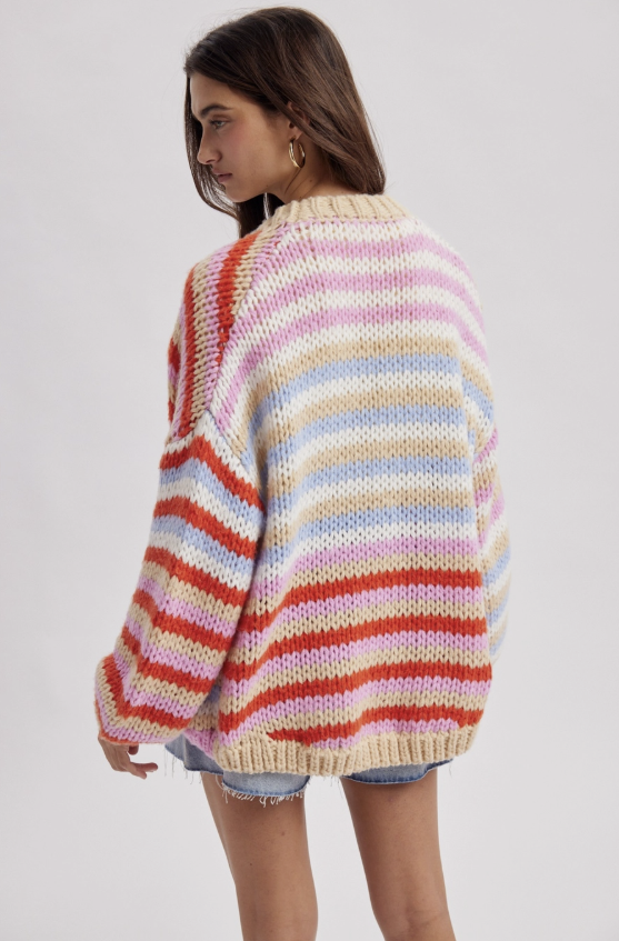 A woman wearing an oversized, chunky-knit striped sweater with horizontal bands of cream, pink, orange, and light blue, shown from a three-quarters back view against a light gray studio background.