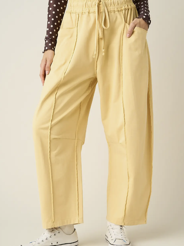 A person wearing pale yellow wide-leg drawstring pants with front patch pockets, styled with a dark polka-dot long-sleeve top and white sneakers against a plain light beige studio background.