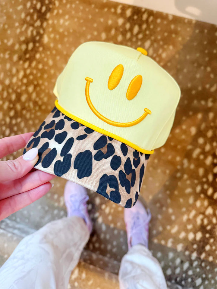 Cap with smiley face design and leopard print brim held by a hand against a patterned background
