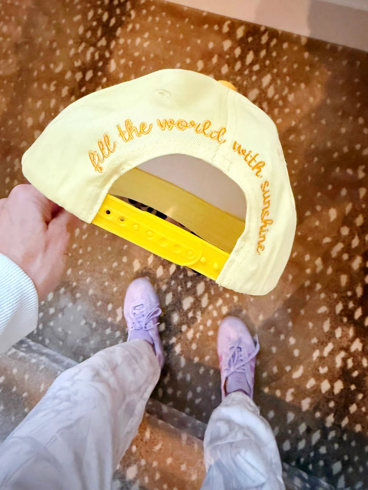 Yellow cap with text held by a person wearing pink shoes on a speckled floor.