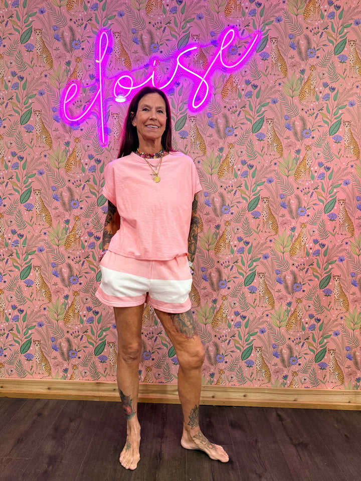 Person in a pink outfit standing against a patterned wall with a neon sign.