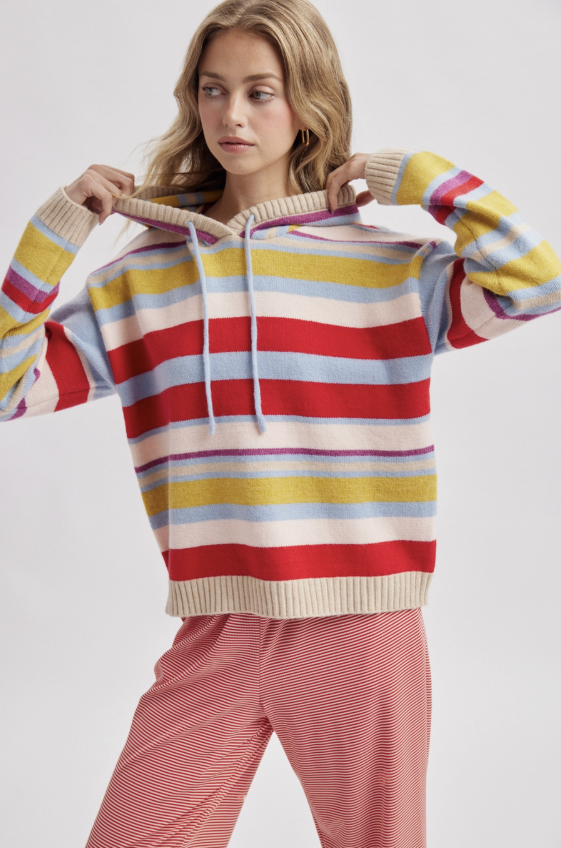 A woman wearing a colorful horizontally striped knit hoodie with red, yellow, light blue, and cream stripes, paired with pink striped pants, against a light gray studio background.