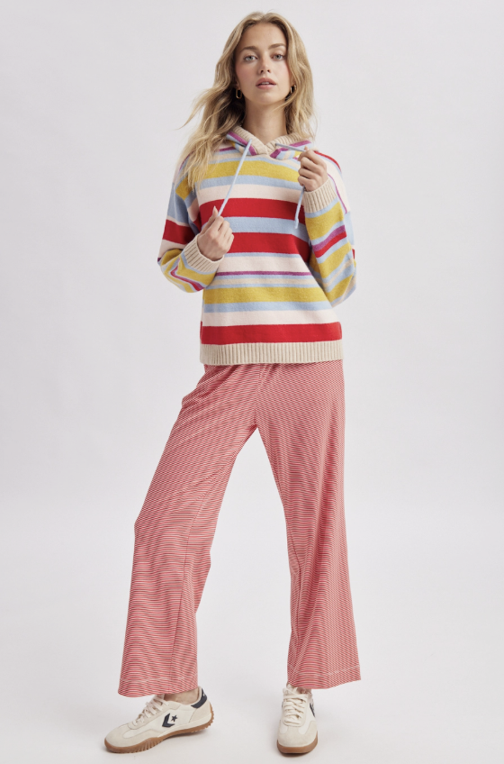 A woman modeling a colorful striped knit hoodie with pink wide-leg pinstripe pants and white sneakers, photographed against a light gray studio background.