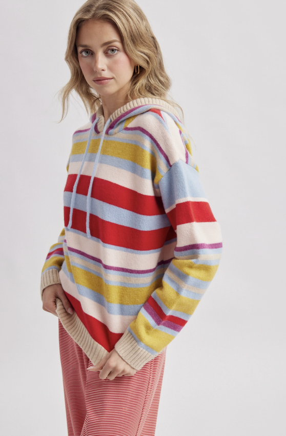 A woman wearing a colorful striped knit hoodie with red, yellow, blue, and cream horizontal stripes, paired with pink striped pants, photographed against a light gray studio background.