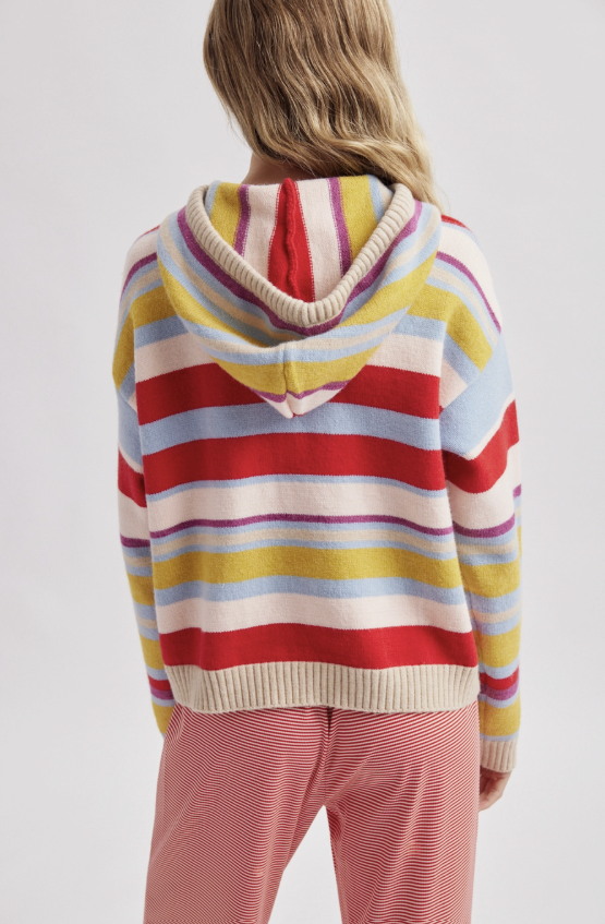 A person shown from the back wearing a colorful striped knit hoodie with horizontal bands of red, yellow, light blue, white, and purple, paired with pink striped pants, against a light gray studio background.