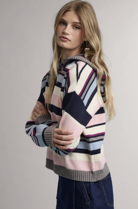 A woman wearing a pastel striped knit sweater with navy, pink, light blue, and cream horizontal blocks, paired with dark blue jeans, photographed against a neutral light gray studio background.