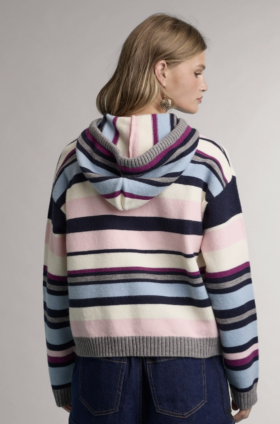 A woman shown from the back wearing a striped knit hoodie with horizontal bands of navy, light blue, pink, cream, and purple, paired with dark blue jeans against a neutral gray studio background.