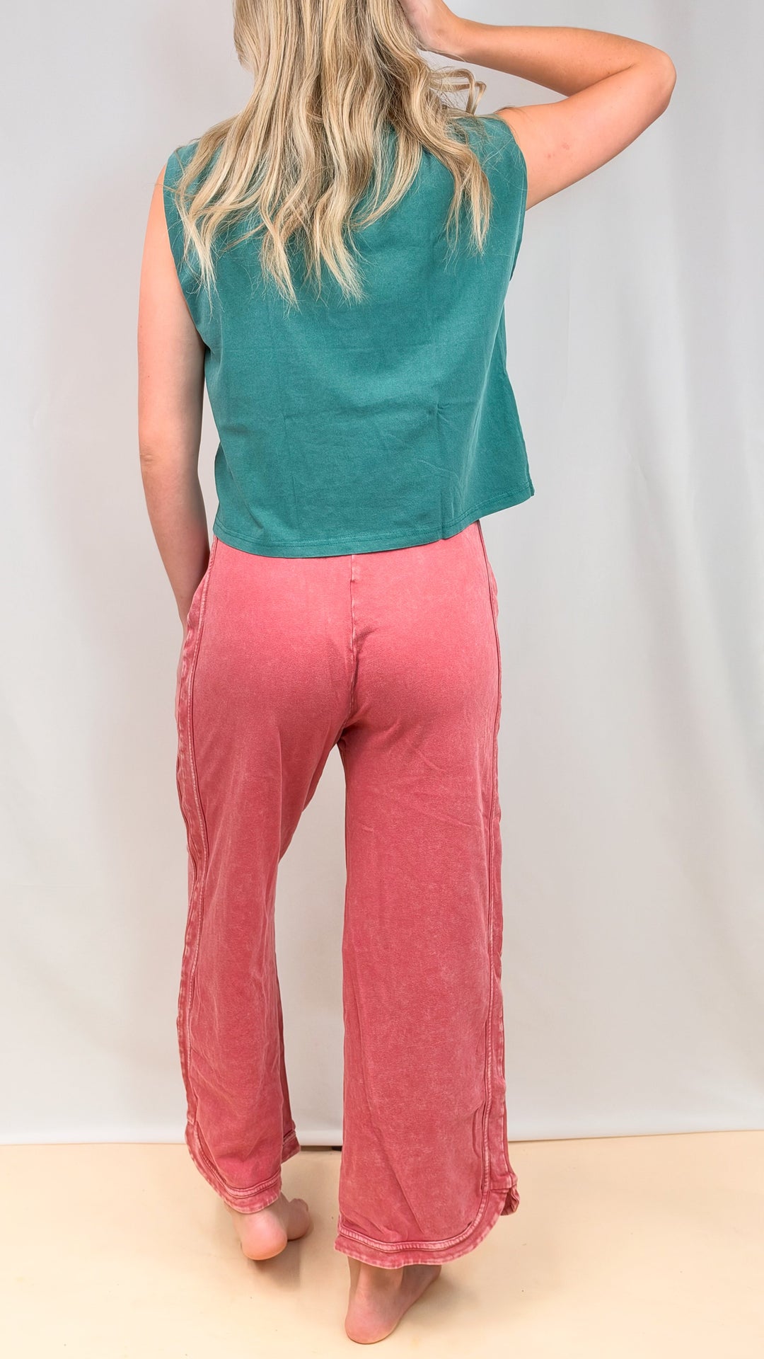 Woman shown from back view wearing teal muscle tee and raspberry color pants on plain background